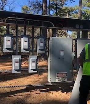 A worker in a high-visibility vest stands near electrical equipment and outdoor disconnect boxes at a solar installation.