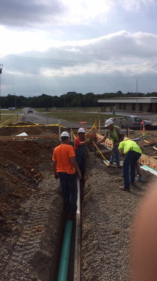 Four workers in hard hats and high-visibility clothing install a pipe in a gravel-filled trench at a construction site.