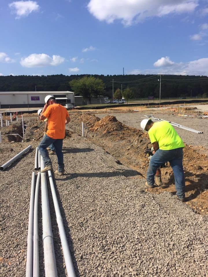 Two construction workers wearing hard hats and high-visibility shirts work in a gravel-covered outdoor site.