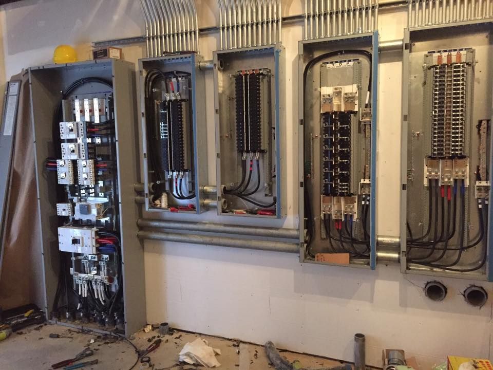 A row of four open electrical breaker panels mounted on a wall with numerous conduit pipes running into the tops.