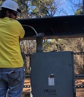 A worker in a hard hat and yellow shirt connects wires to a Prism Electric equipment box in a wooded area.