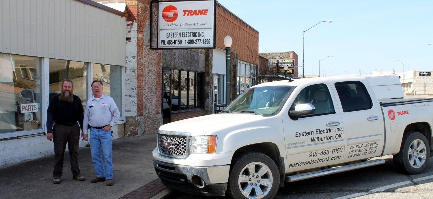 Two men stand in front of a storefront and a parked white pickup truck with logo branding in a sunny outdoor setting.