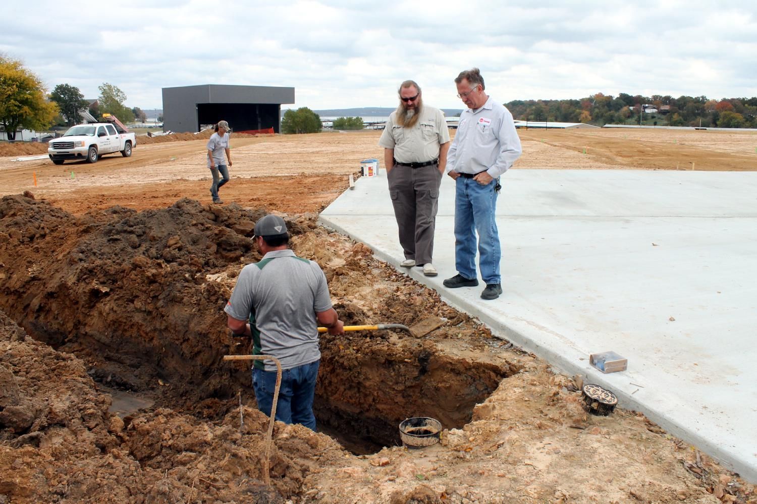 Three people observe a construction site with a worker digging a trench beside a concrete slab foundation.