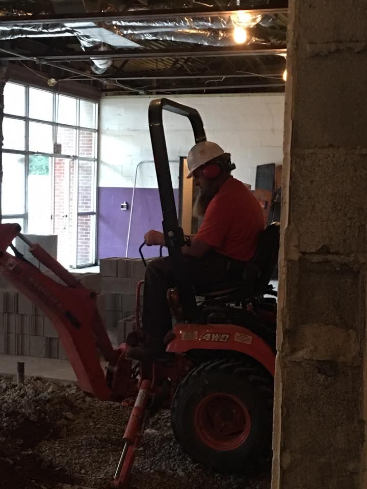 A person wearing a white hard hat operates a red backhoe indoors on a dirt floor near a concrete wall.