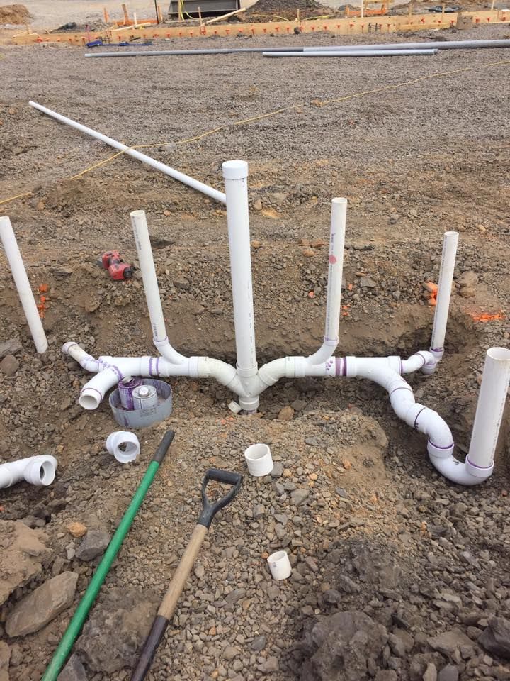 White PVC plumbing pipes for a construction project, arranged in a network with vertical risers in a dirt trench.