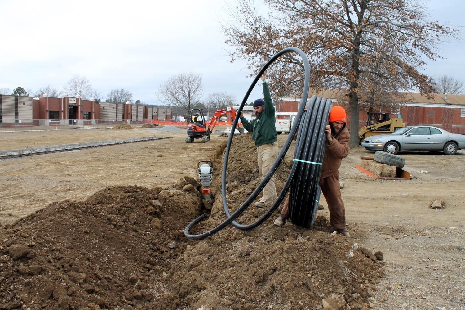 Two workers install a large, coiled black corrugated pipe into an open trench on an outdoor construction site.