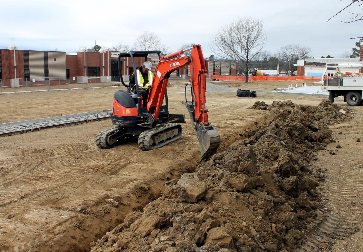 An orange mini-excavator digs a trench in an open dirt lot near a brick building.