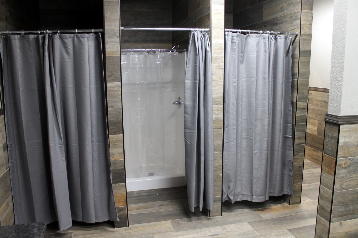 Three shower stalls with grey curtains, wood-look tiled walls, and light flooring in a restroom.