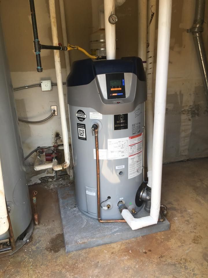 A gray residential water heater sits on a concrete pad with white plumbing pipes and electrical components.