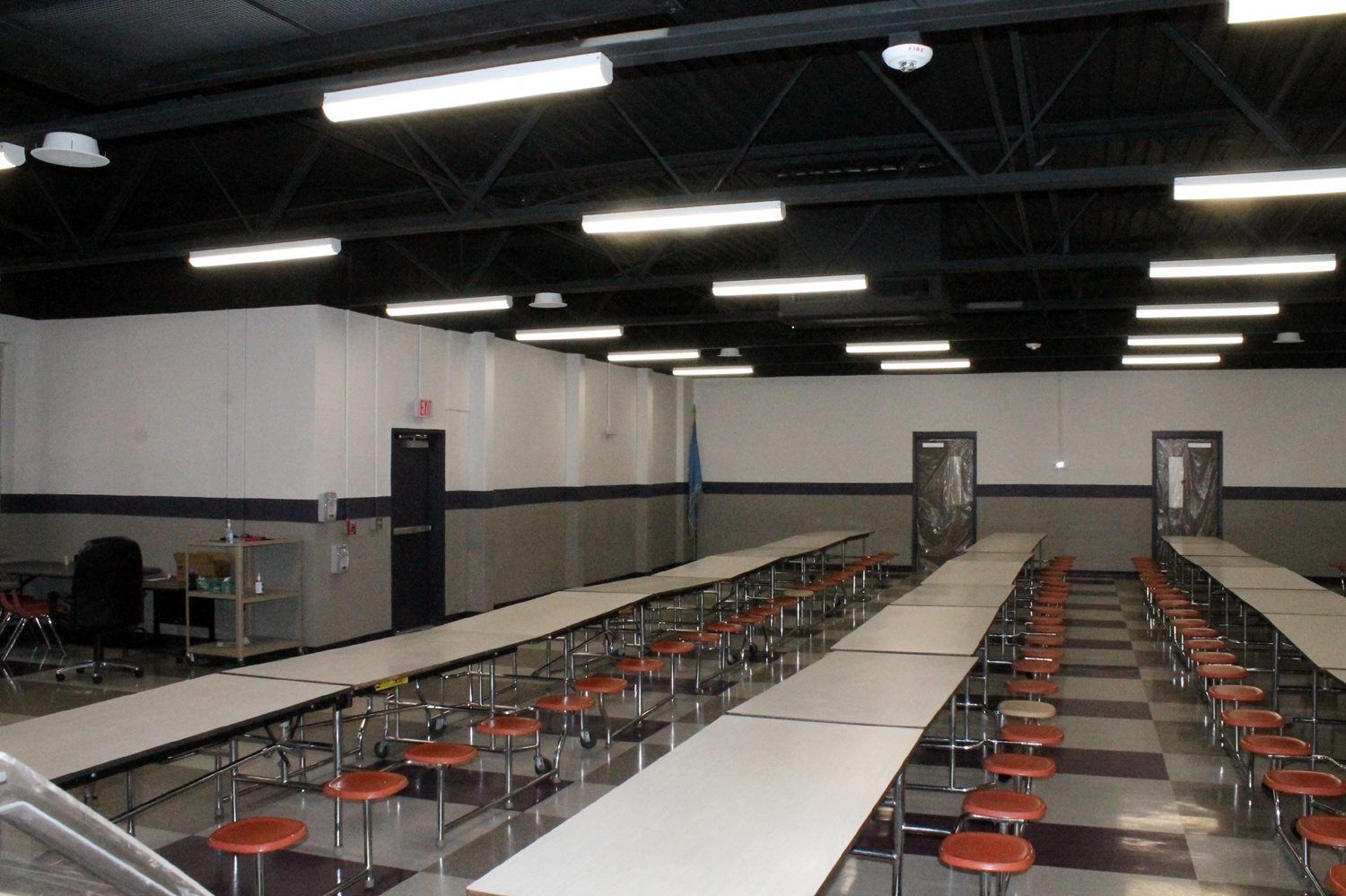 A cafeteria with rows of long tables and attached orange stools under a black industrial ceiling with fluorescent lights.