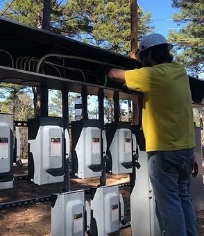 A worker in a yellow shirt and hard hat installs wiring in outdoor electrical meter boxes.