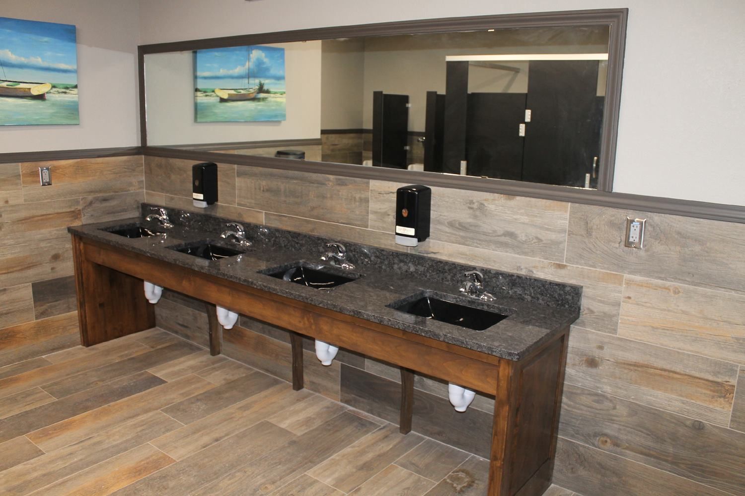A public restroom featuring a long vanity with dark stone countertops, three sinks, soap dispensers, and a large mirror.