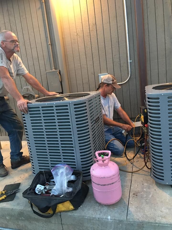 Two technicians service two outdoor HVAC units on a concrete patio, using a pink refrigerant tank and tool bag.