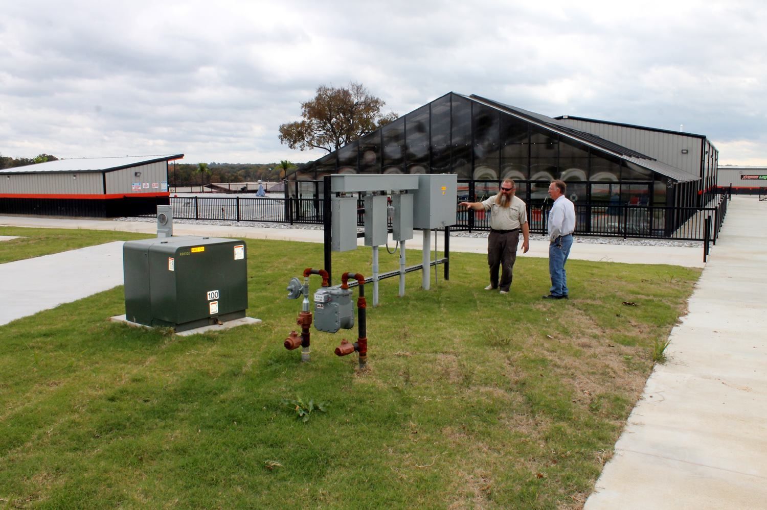 Two people stand on a grassy lawn near an outdoor utility installation and a large modern building with glass walls.