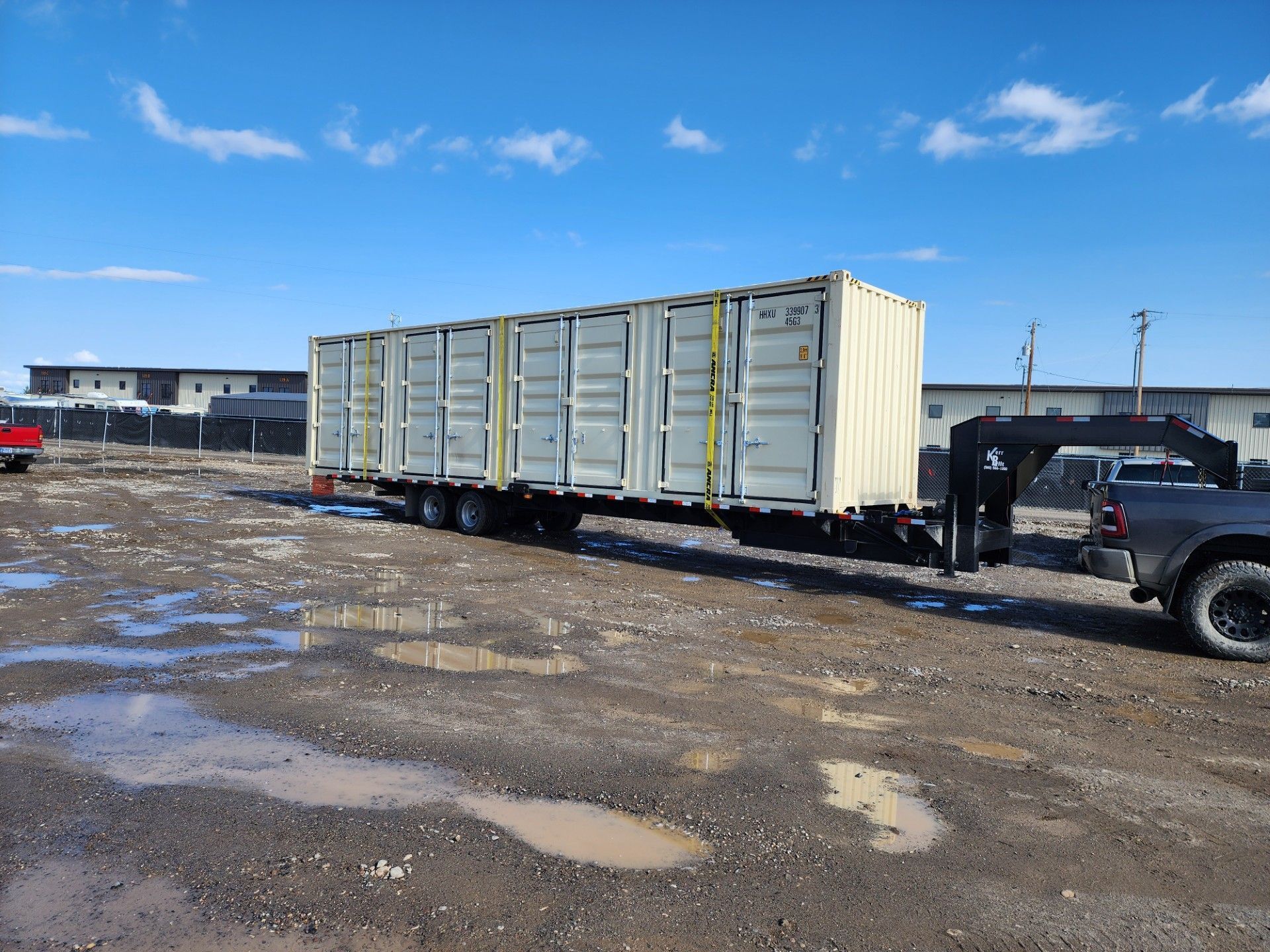 A truck towing a long, white shipping container on a trailer across a muddy gravel lot under a blue sky.