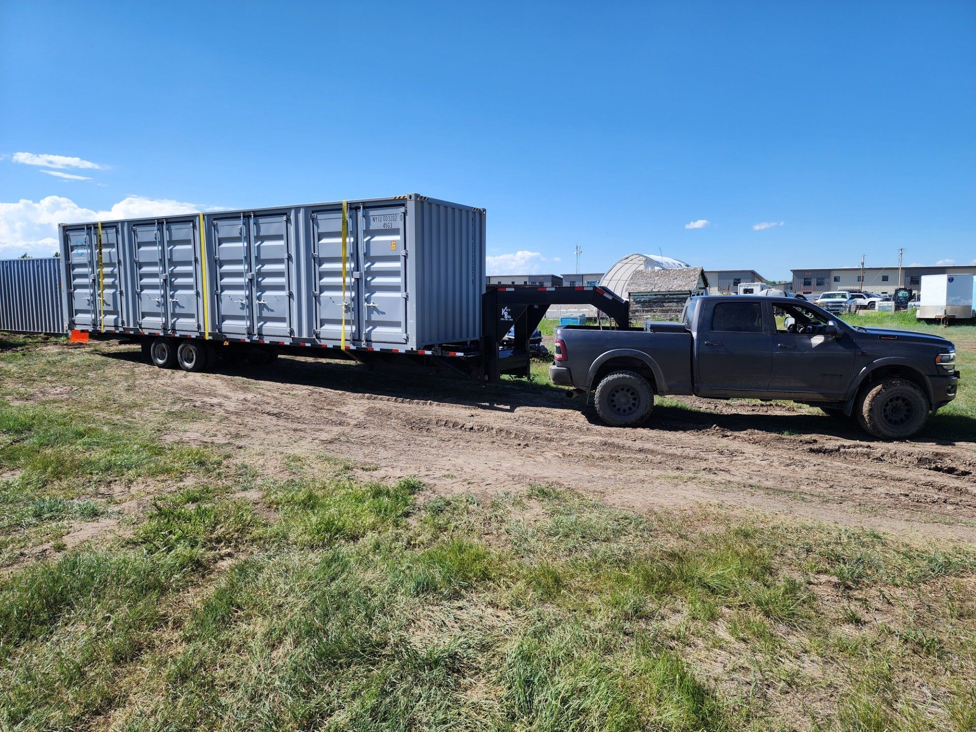 Dark pickup truck towing a long trailer holding multiple shipping containers on a grassy field.
