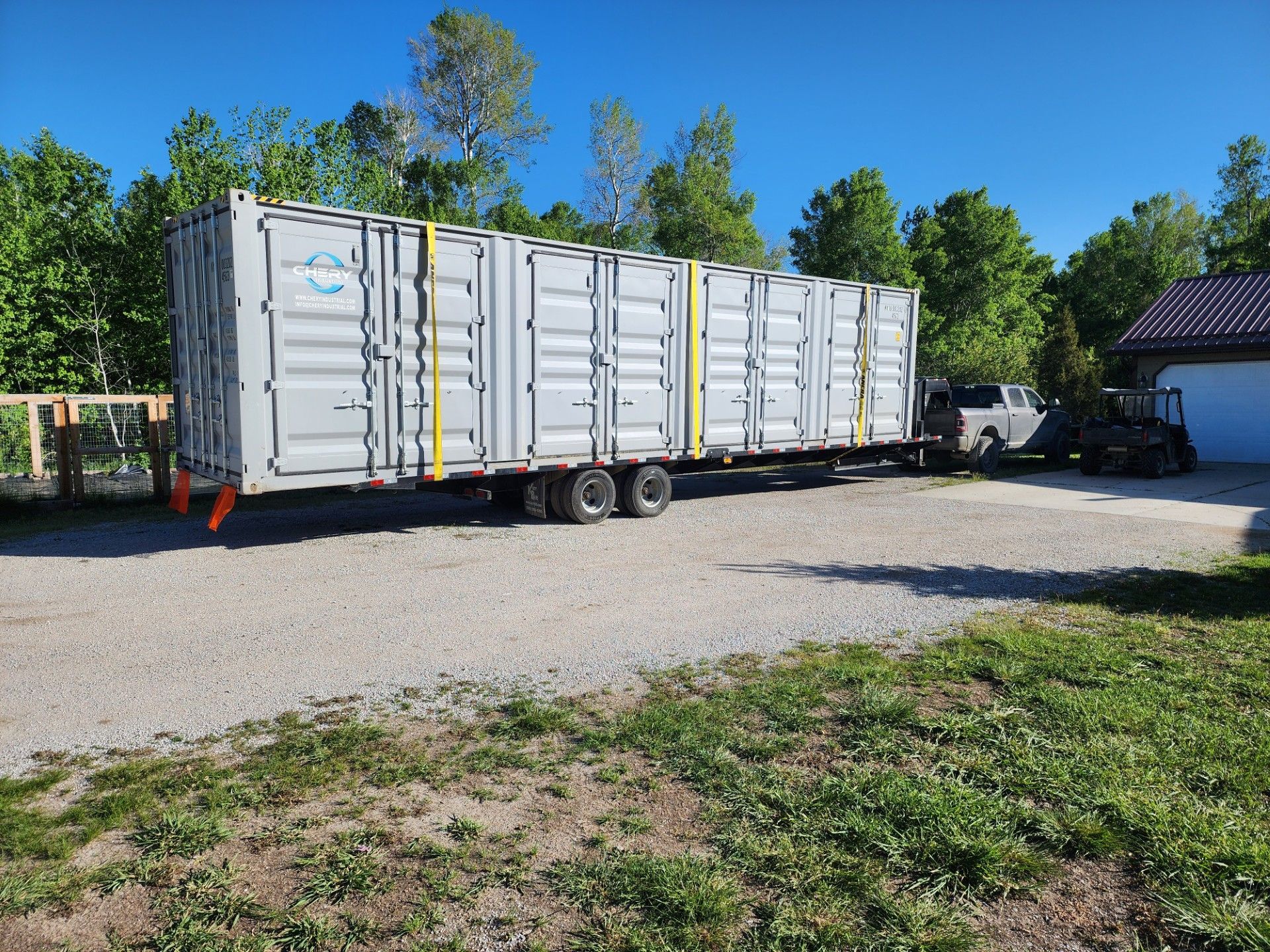 Gray shipping containers loaded on a trailer on a gravel driveway, blue sky, green trees.