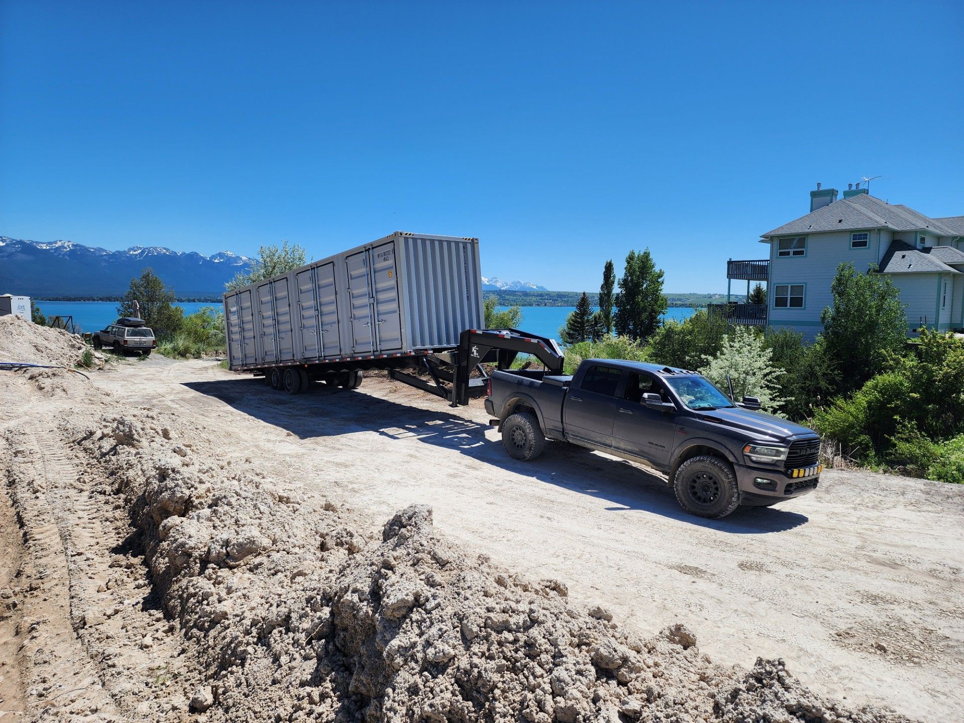 Truck pulling a large shipping container on a muddy road near a house with mountains in the background.