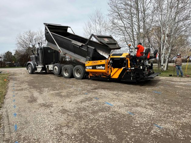 Orange paving machine loading black asphalt into a dump truck on a gravel lot, with workers nearby