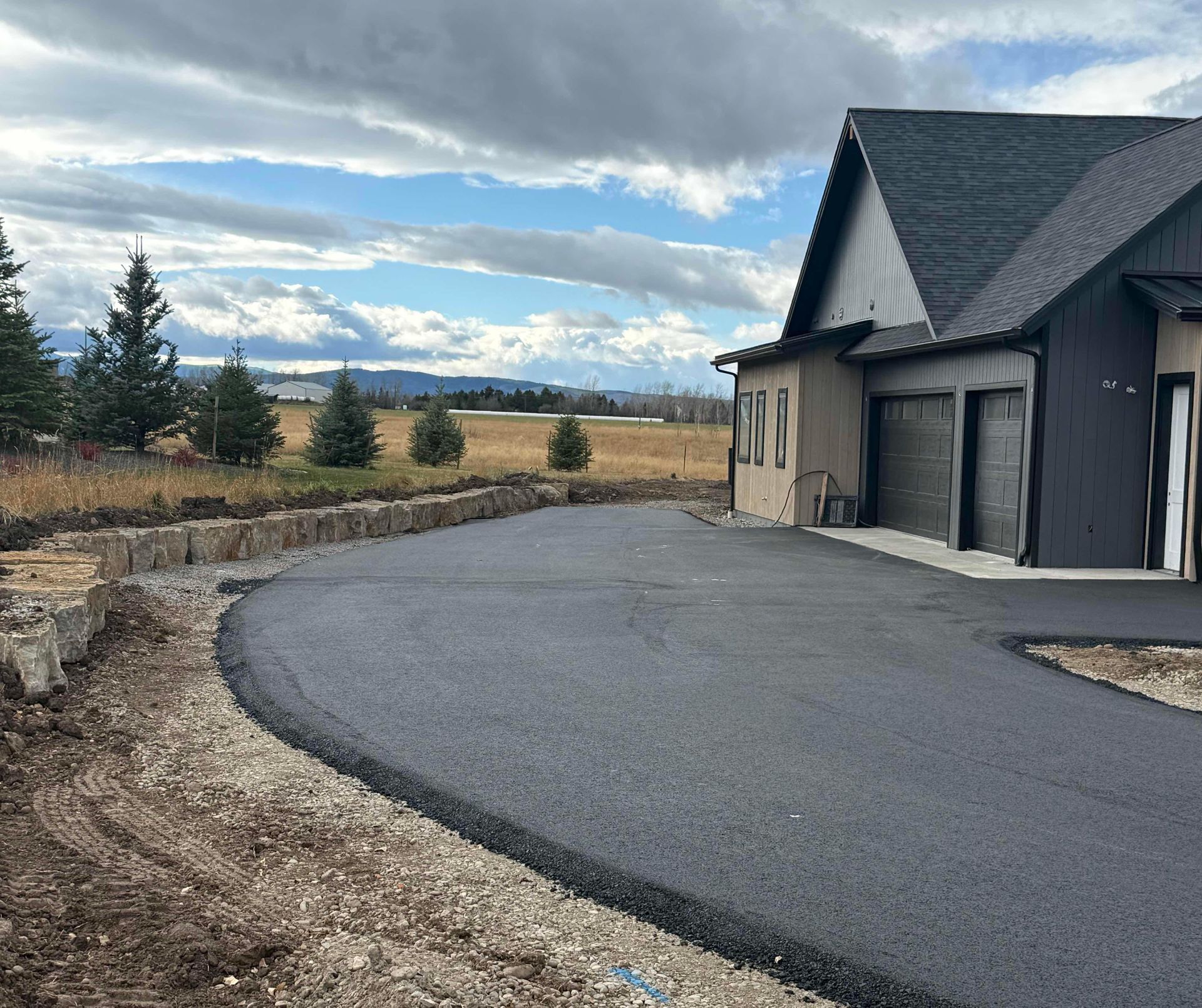 Curved gravel driveway beside a dark house with garage, overlooking open fields under a cloudy sky