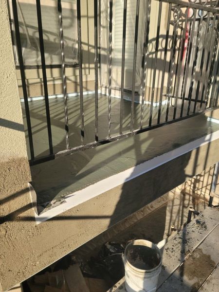 A beige balcony ledge with a black metal railing, featuring a white taped edge and a bucket sitting on the ground below.