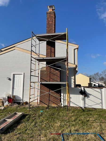 Metal scaffolding stands against a brick chimney on the side of a light-colored, two-story house under a blue sky.