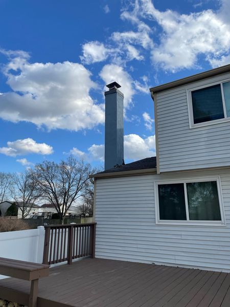 Exterior view of a house with white siding and a deck, featuring a tall, gray chimney against a blue, cloudy sky.