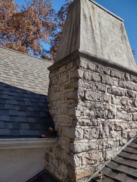 A stone chimney rises above a shingled roof, with visible debris accumulation where the chimney meets the roofline.