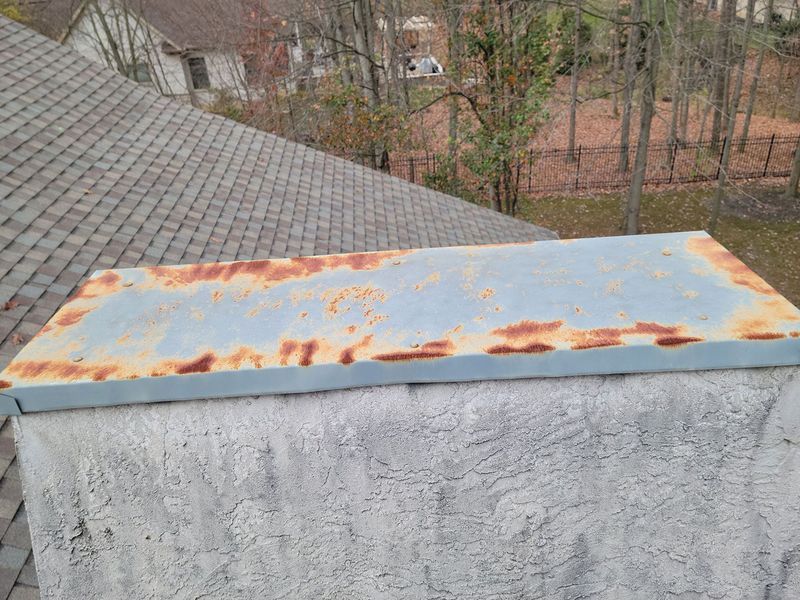 A weathered, light blue metal chimney cap with significant rust along the edges, situated on a textured masonry chimney.