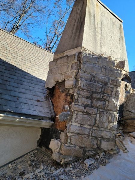 A stone chimney on a roof has collapsed, revealing the wooden structure underneath and debris scattered on the shingles.