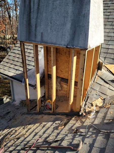 A chimney structure on a shingled roof with its exterior siding removed, revealing wooden wall framing and interior plywood.