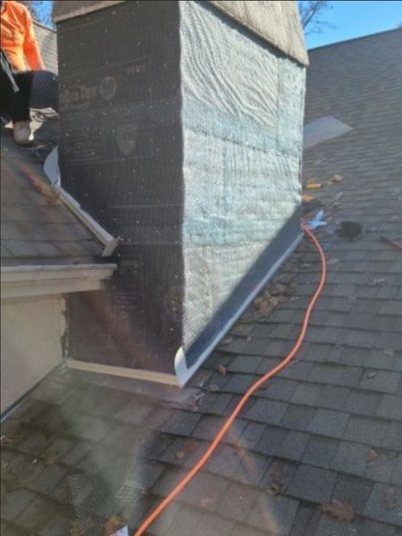 A worker stands on a roof next to a chimney covered in black roofing felt and white insulation underlayment.