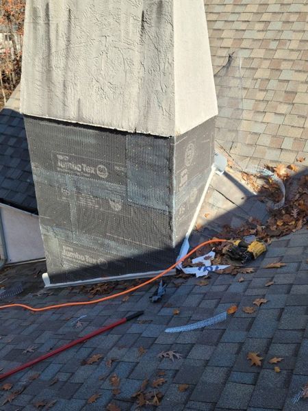 A chimney exterior under repair, featuring exposed grey protective sheathing and a beige upper section on a shingled roof.