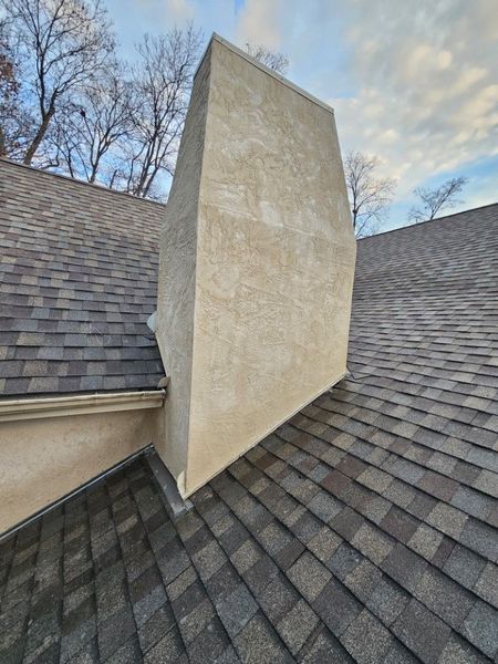 A beige stucco chimney rises from a brown shingled roof under a clear, late afternoon sky.