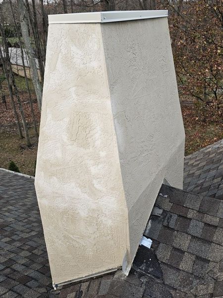 A beige, stucco-finished chimney shroud sits on an asphalt shingle roof.