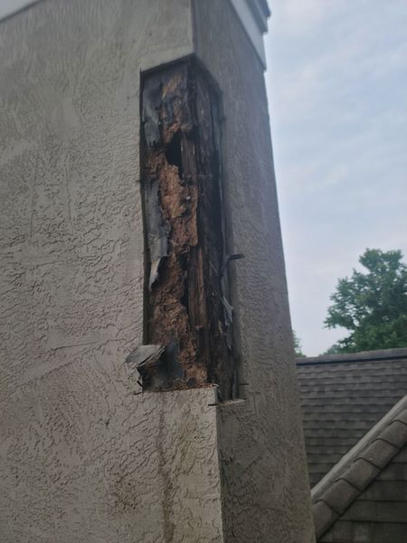 A vertical, textured stucco exterior wall section has a large, rectangular opening exposing severely rotted wooden framing.