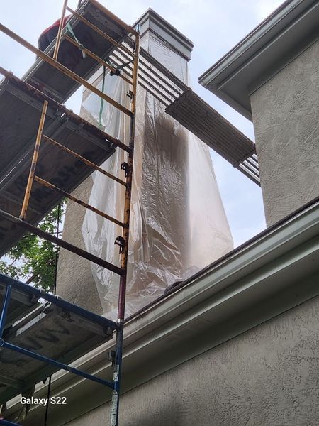 A tall chimney covered in plastic sheeting stands behind construction scaffolding next to a stucco house.