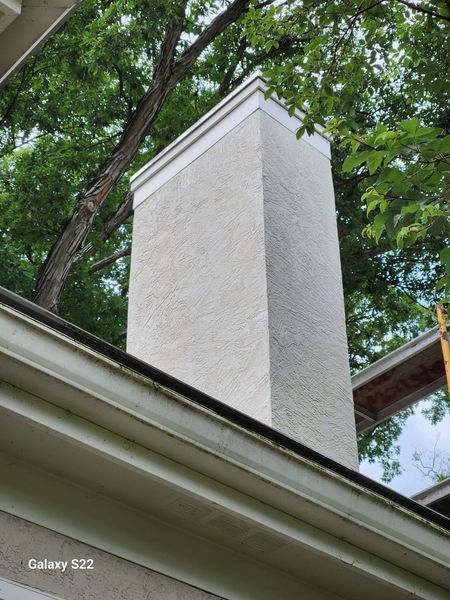 A white, stucco-finished chimney rises from a home’s roofline surrounded by green tree branches.