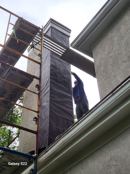 A construction worker stands on scaffolding, applying a black moisture barrier to the side of a tall chimney.