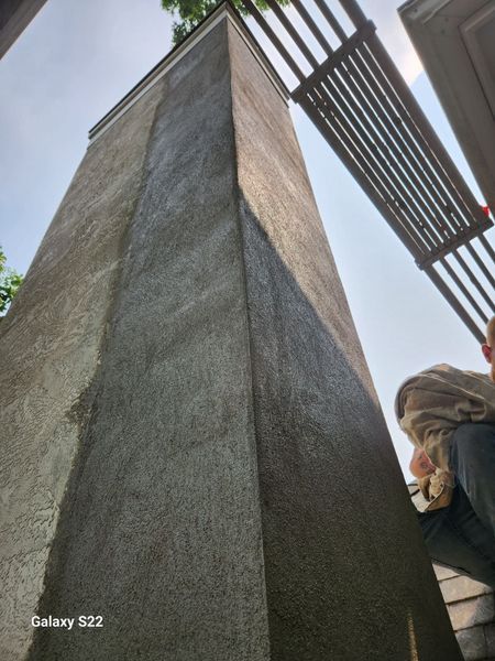 A tall, textured stucco pillar stands outdoors next to a slatted wooden pergola.