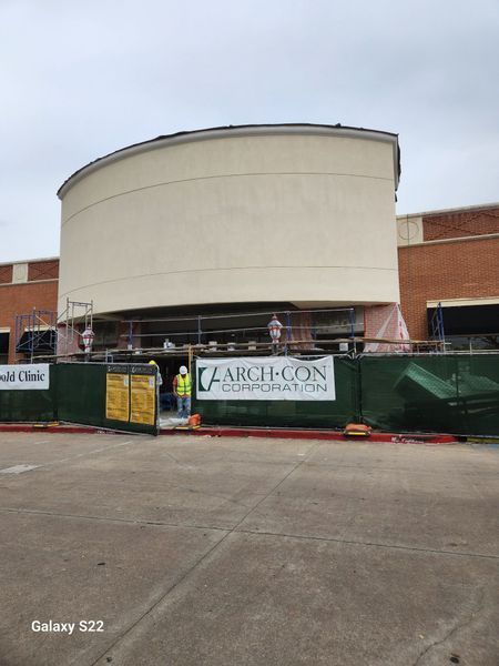 A commercial building under construction with a large curved facade, scaffolding, and a green construction fence.