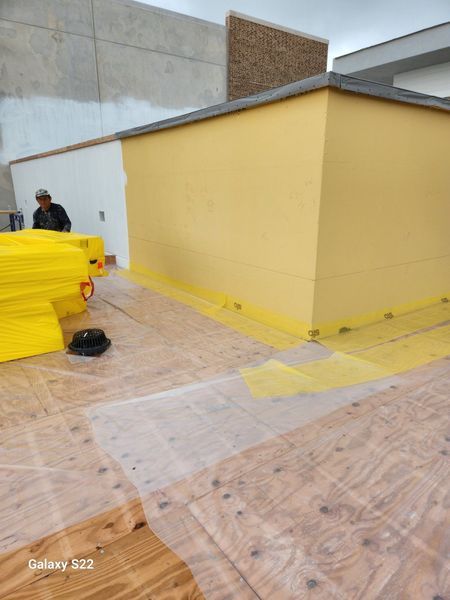 A worker paints a yellow wall on a rooftop, with plywood flooring and white plastic sheeting laid out nearby.