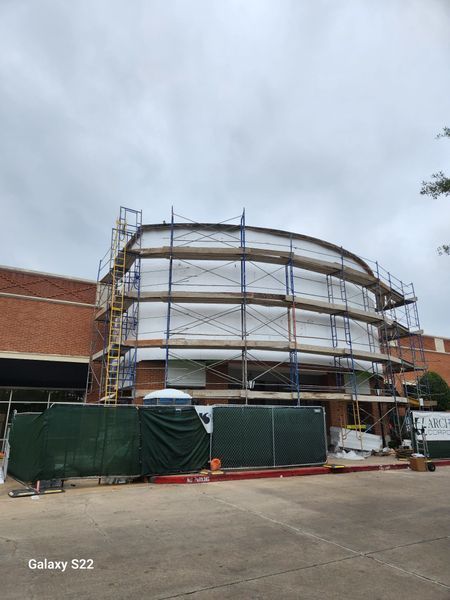 Scaffolding surrounds the curved brick exterior of a building under renovation, partially blocked by a green privacy fence.