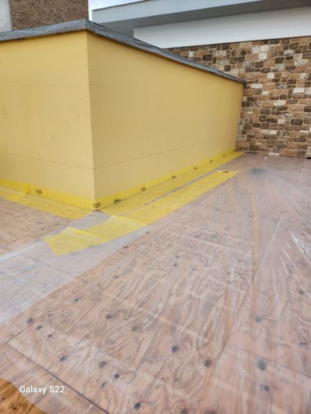 Construction site showing a yellow wall edge with protective yellow tape and plastic sheeting covering the plywood floor.