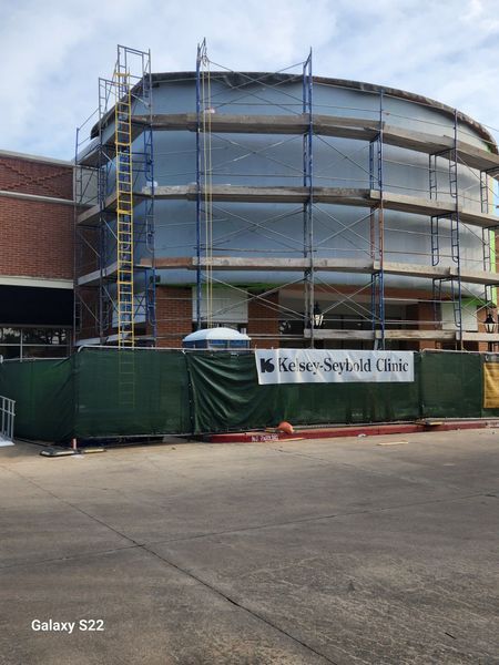 A circular building under construction at Kelsey-Seybold Clinic, surrounded by scaffolding and a green privacy fence.