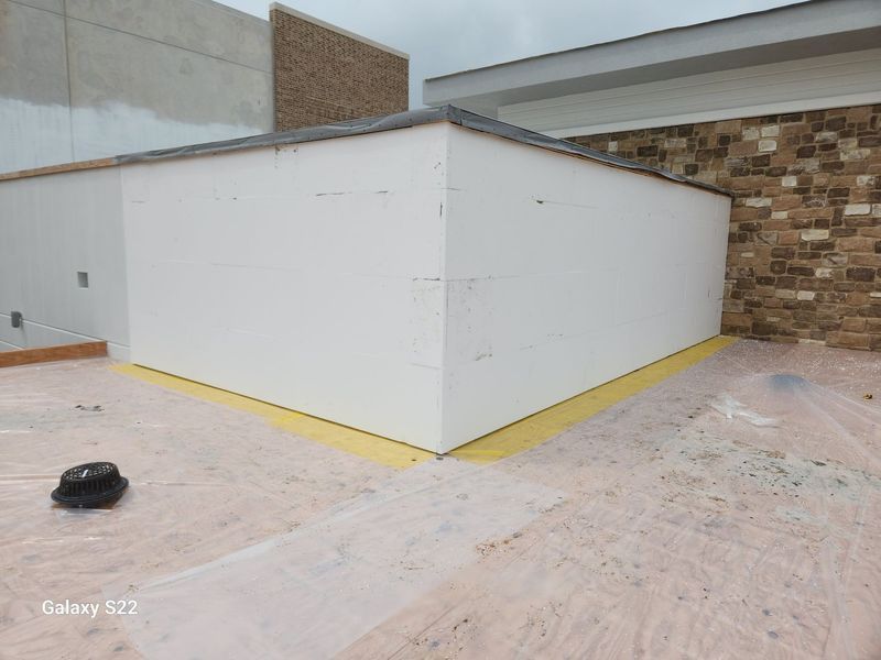 A white masonry corner wall stands on a rooftop covered in light-colored protective plastic, next to a stone-clad wall.