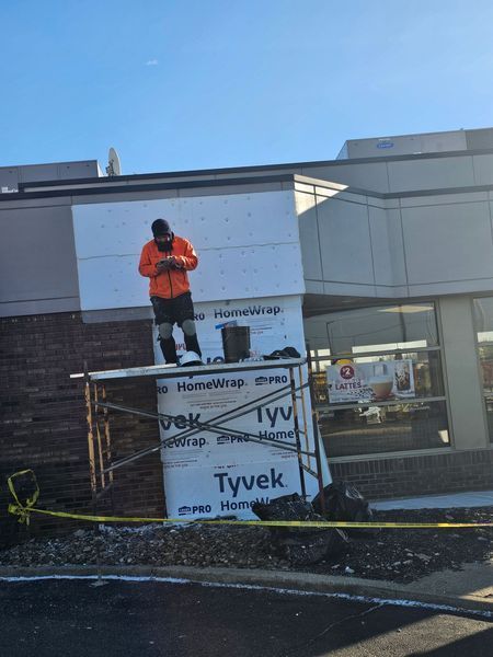A worker in an orange jacket stands on scaffolding in front of a building covered in Tyvek wrap, looking at a mobile phone.