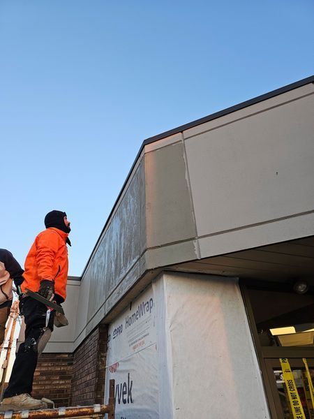 A worker in an orange jacket stands on a platform, installing exterior panels on a building under construction.