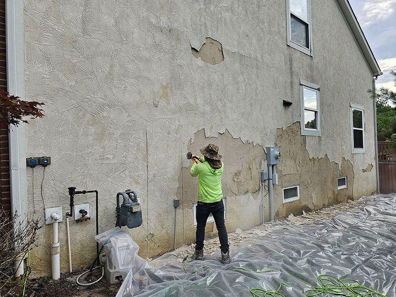 A person in a neon yellow shirt uses a hammer to remove damaged stucco from the side of a house.