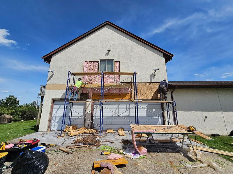 Two workers use scaffolding to remove siding and insulation from the exterior wall above a residential garage.